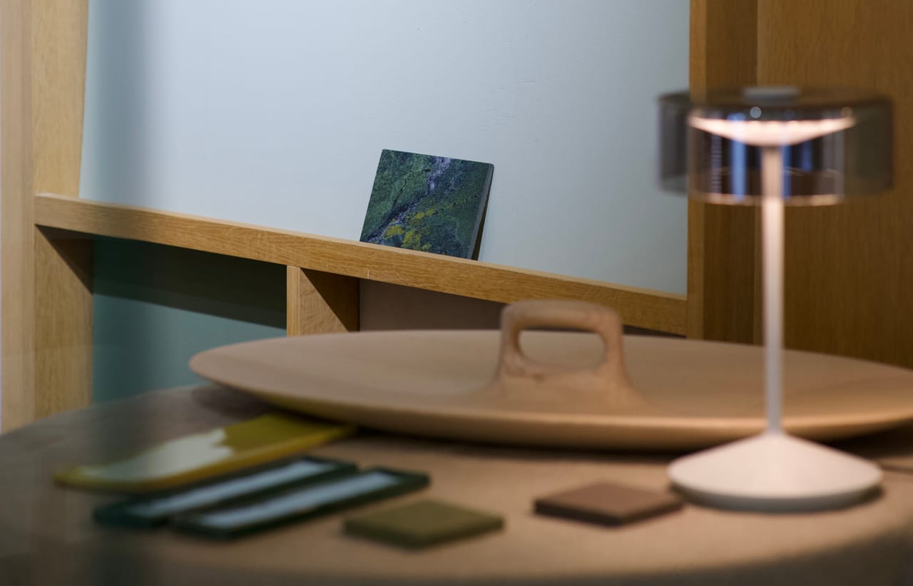 A turned wooden tray on a table with glass, tile and leather samples, a cordless lamp beside it and a stone sample on the shelf behind